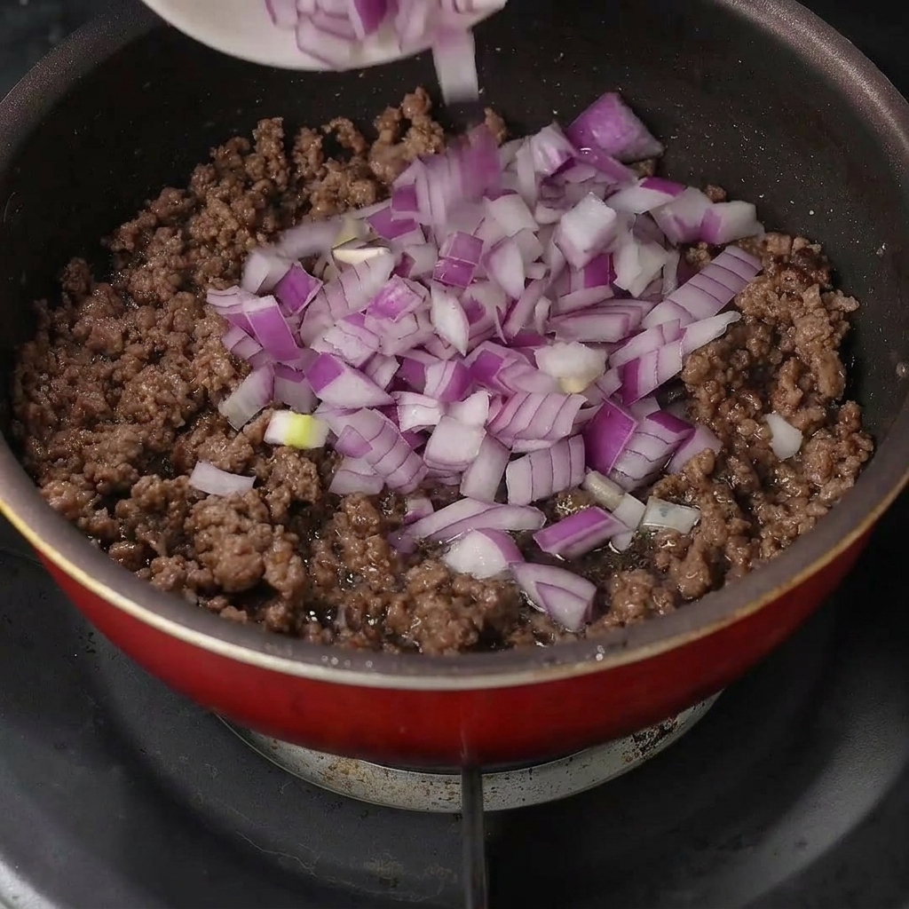 Cebolla morada fresca picada vertida sobre la carne picada cocinada en una sartén.