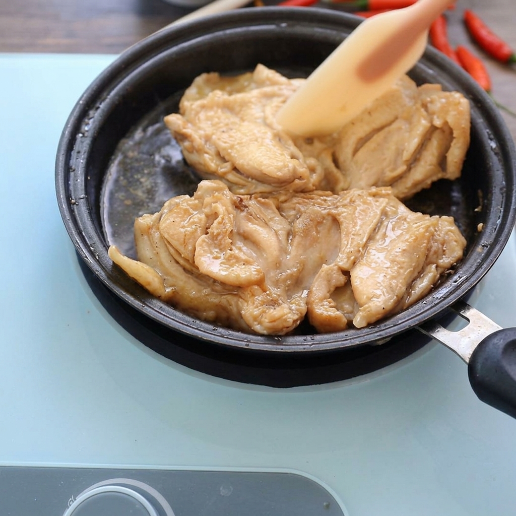 Muslos de pollo cocinados a fuego lento en una sartén con salsa oscura, siendo presionados ligeramente con una espátula de silicona.