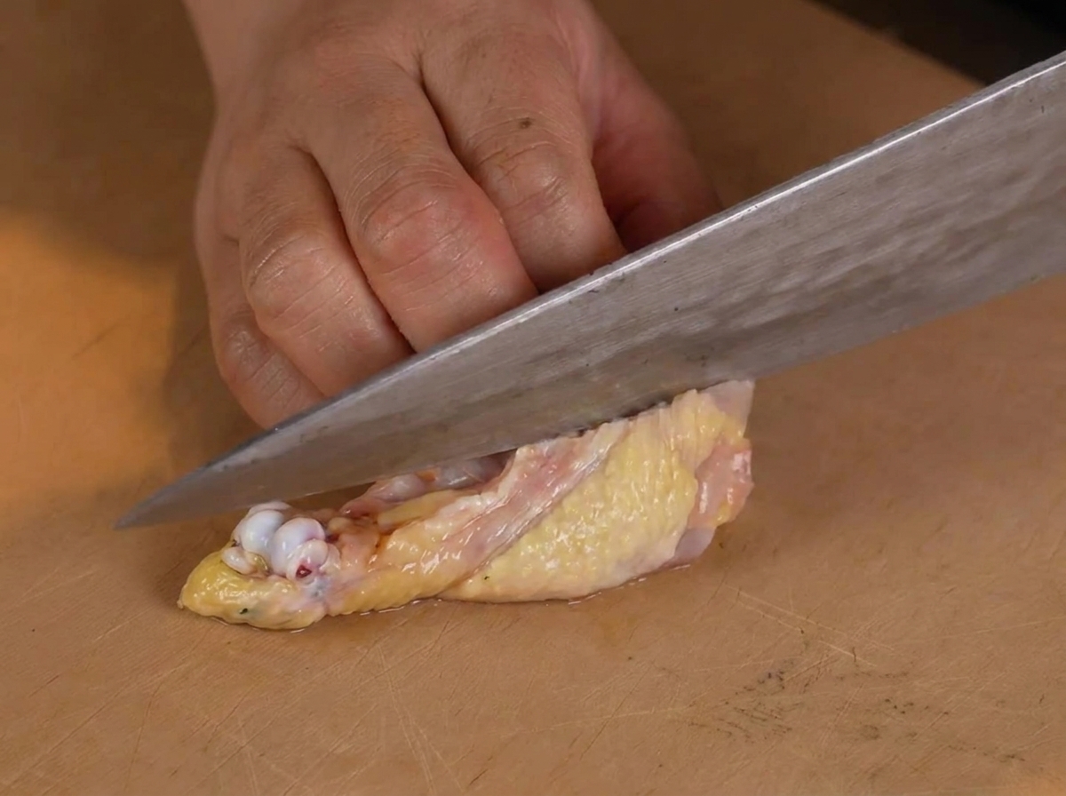 Una persona usando un cuchillo de chef para quitar cuidadosamente el hueso de una pierna de pollo cruda sobre una tabla de madera.