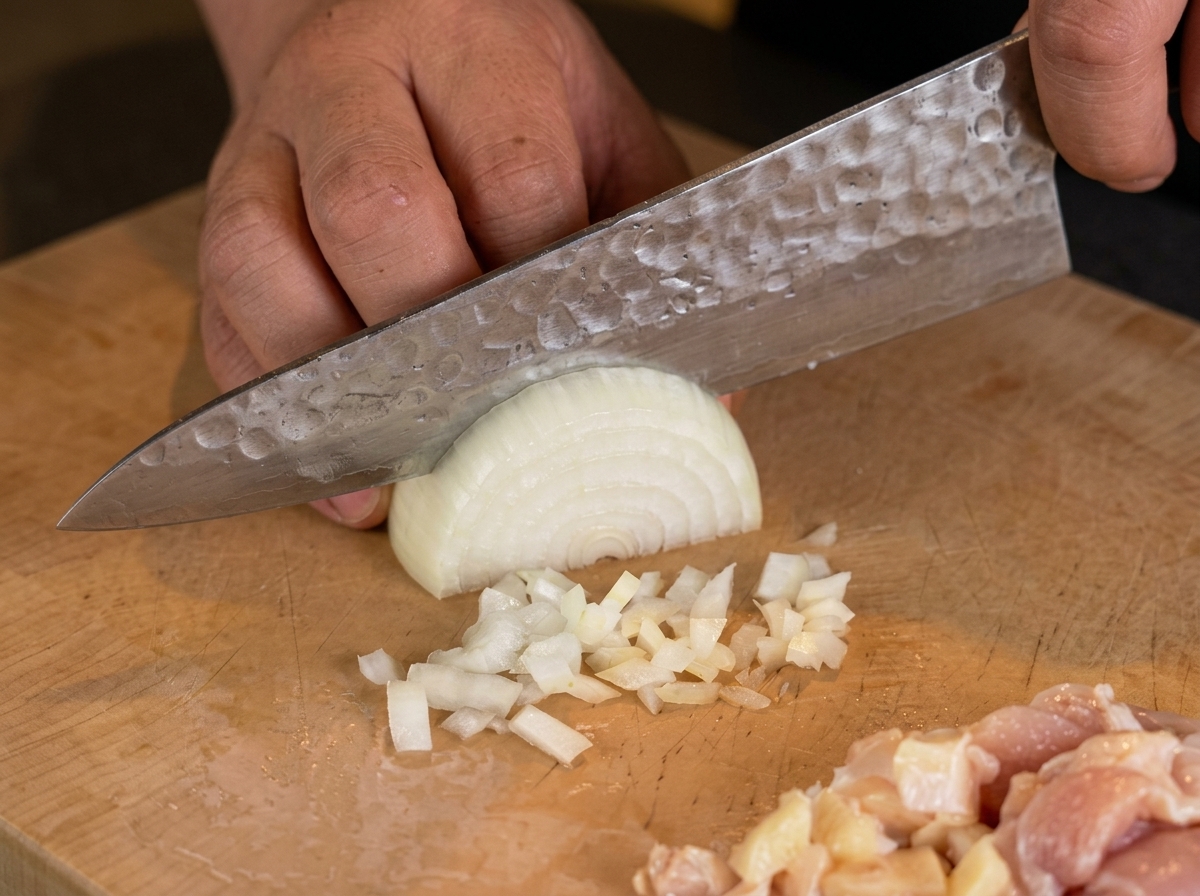 La mano de un chef usando un cuchillo para picar finamente media cebolla blanca sobre una tabla.