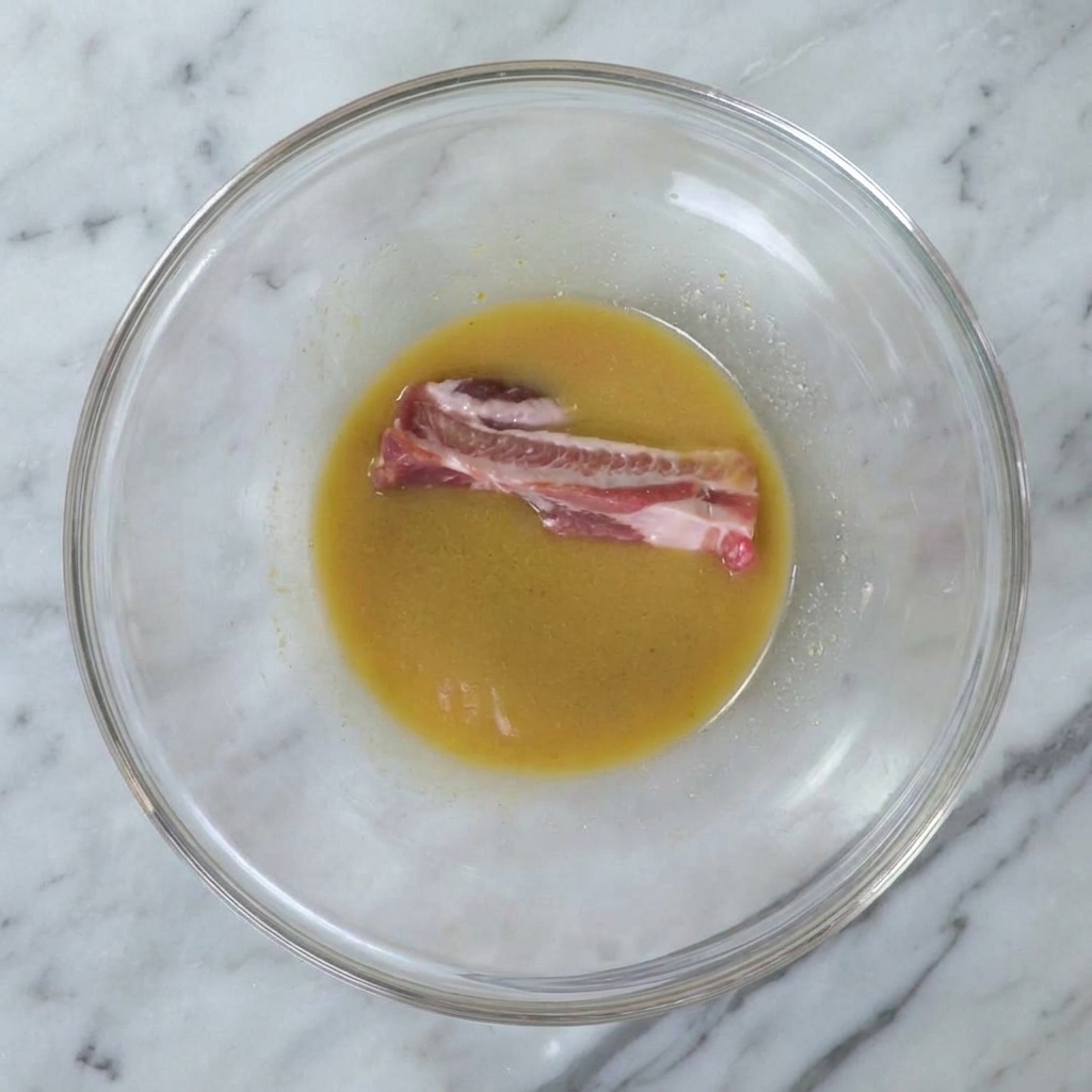 Una costilla cruda siendo sumergida en el marinado de miso dentro de un cuenco grande.