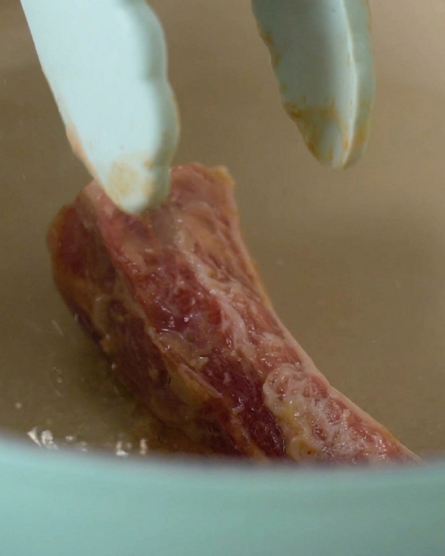 Pinzas colocando una pieza de costilla marinada en la olla caliente.