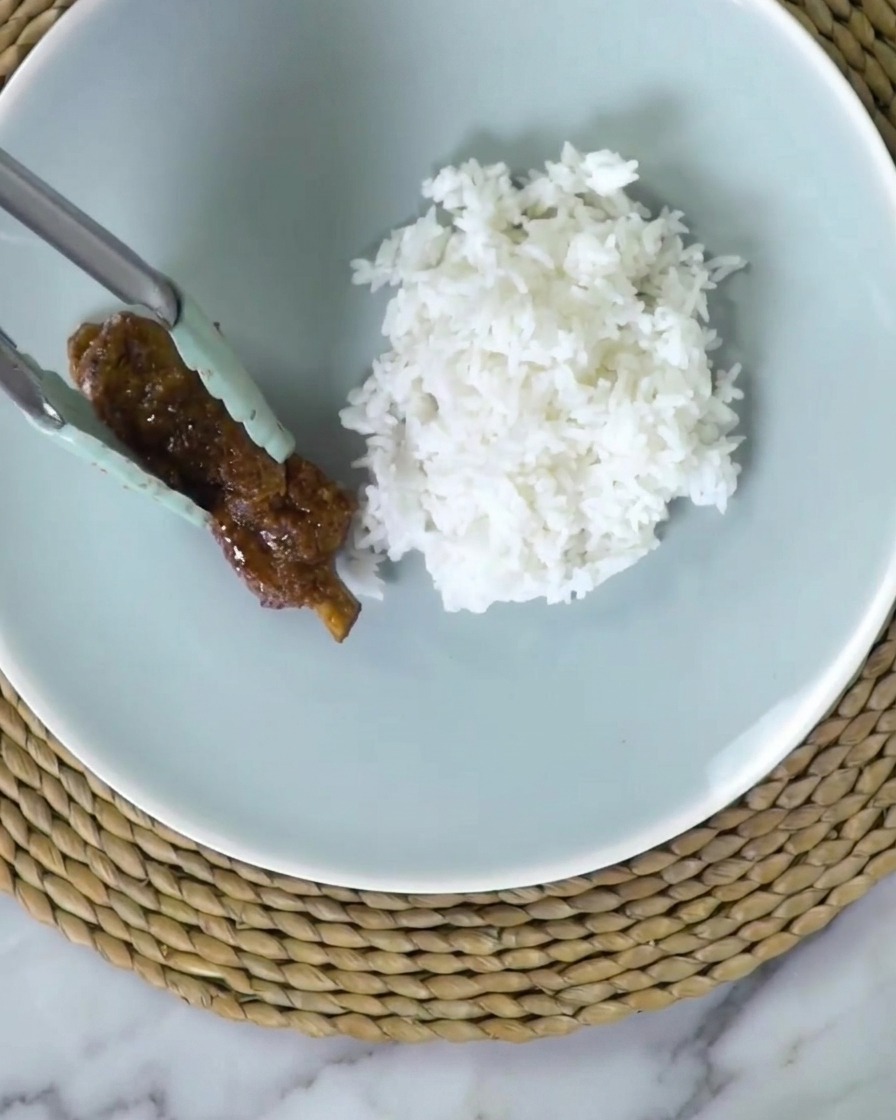 Pinzas colocando una costilla glaseada en un plato junto a arroz blanco.