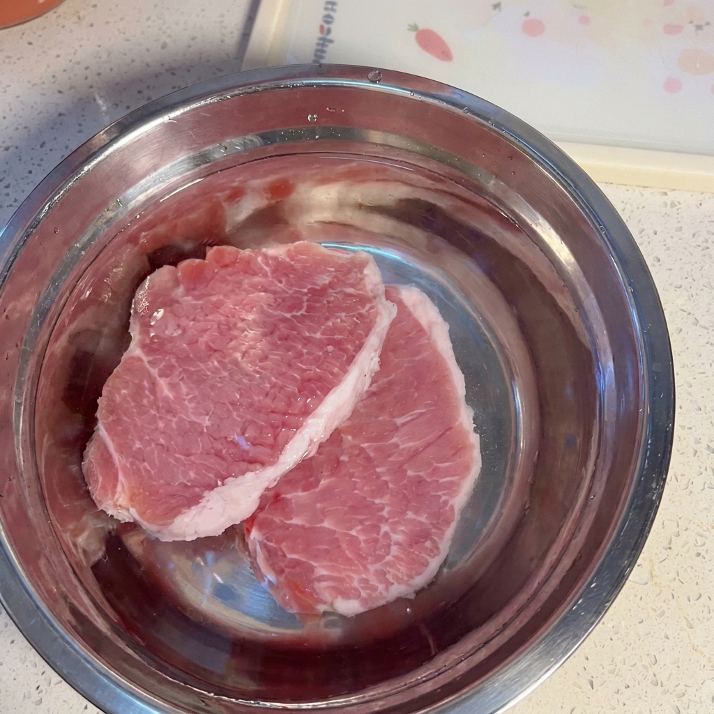 Rodajas de cerdo crudo en un cuenco metálico lleno de agua.
