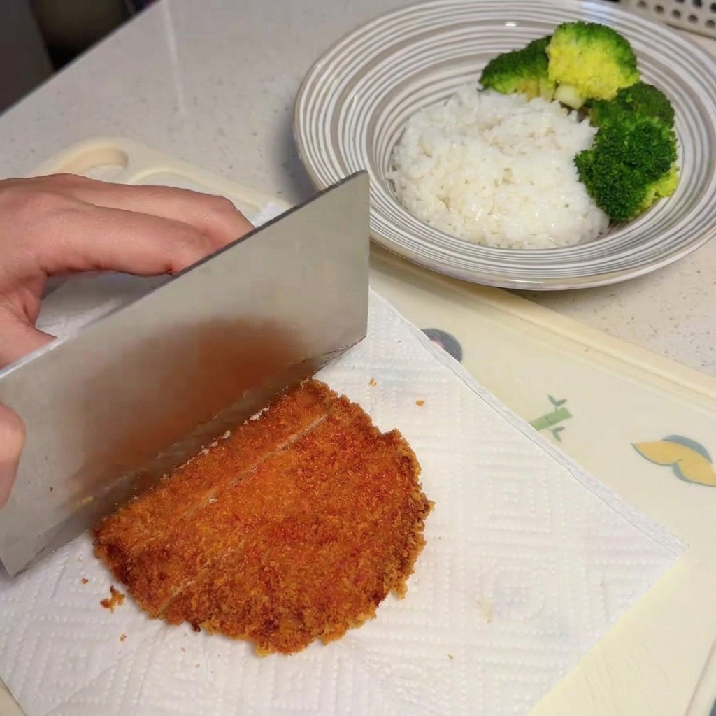 Una mano usando un cuchillo grande para cortar una chuleta dorada en tiras sobre papel absorbente.
