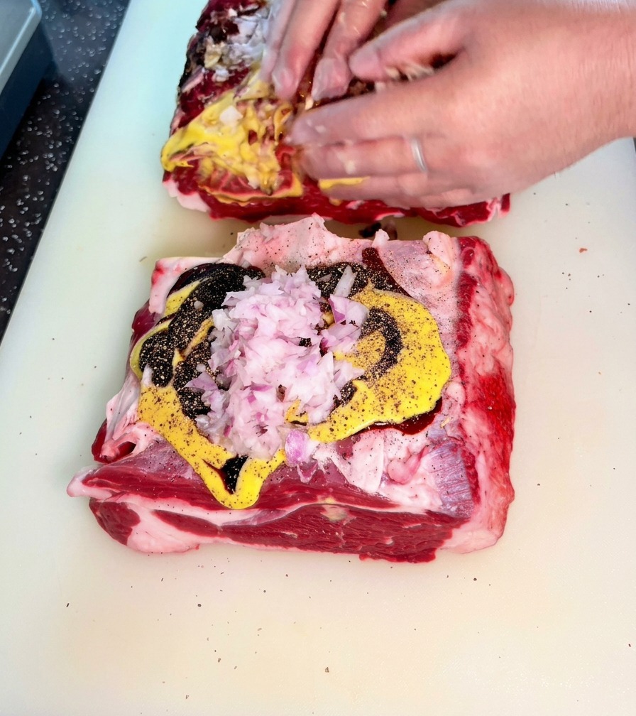 Manos frotando activamente la mezcla de cebollas, mostaza y salsas oscuras en la carne de ternera cruda.