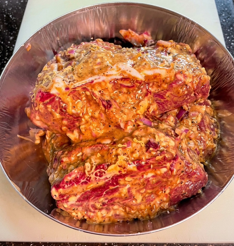 Primer plano de trozos de ternera en un bol de metal, cubiertos por un marinado espeso con trozos visibles de cebolla roja y pimienta.