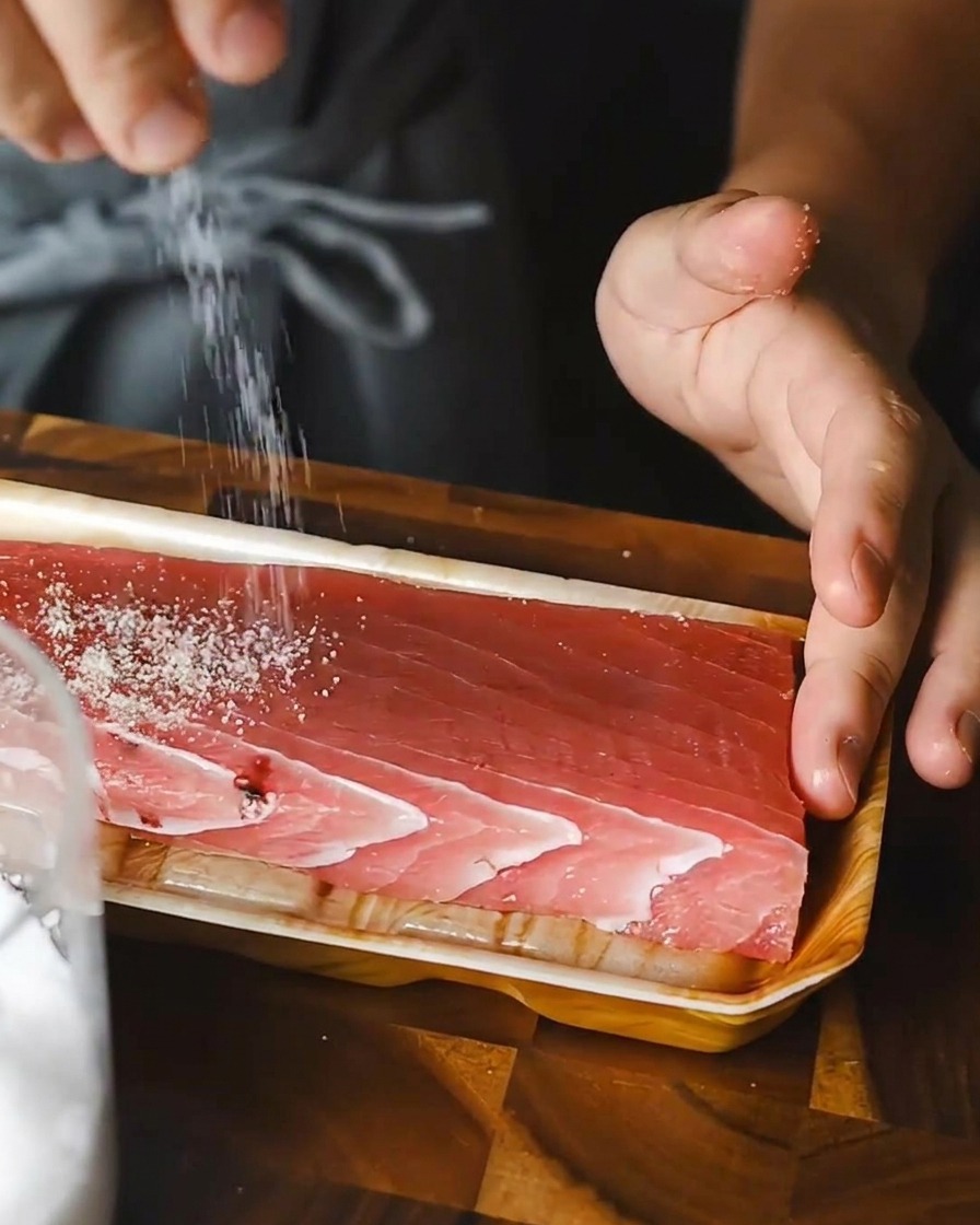 Persona esparciendo sal sobre un bloque rectangular de atún crudo.
