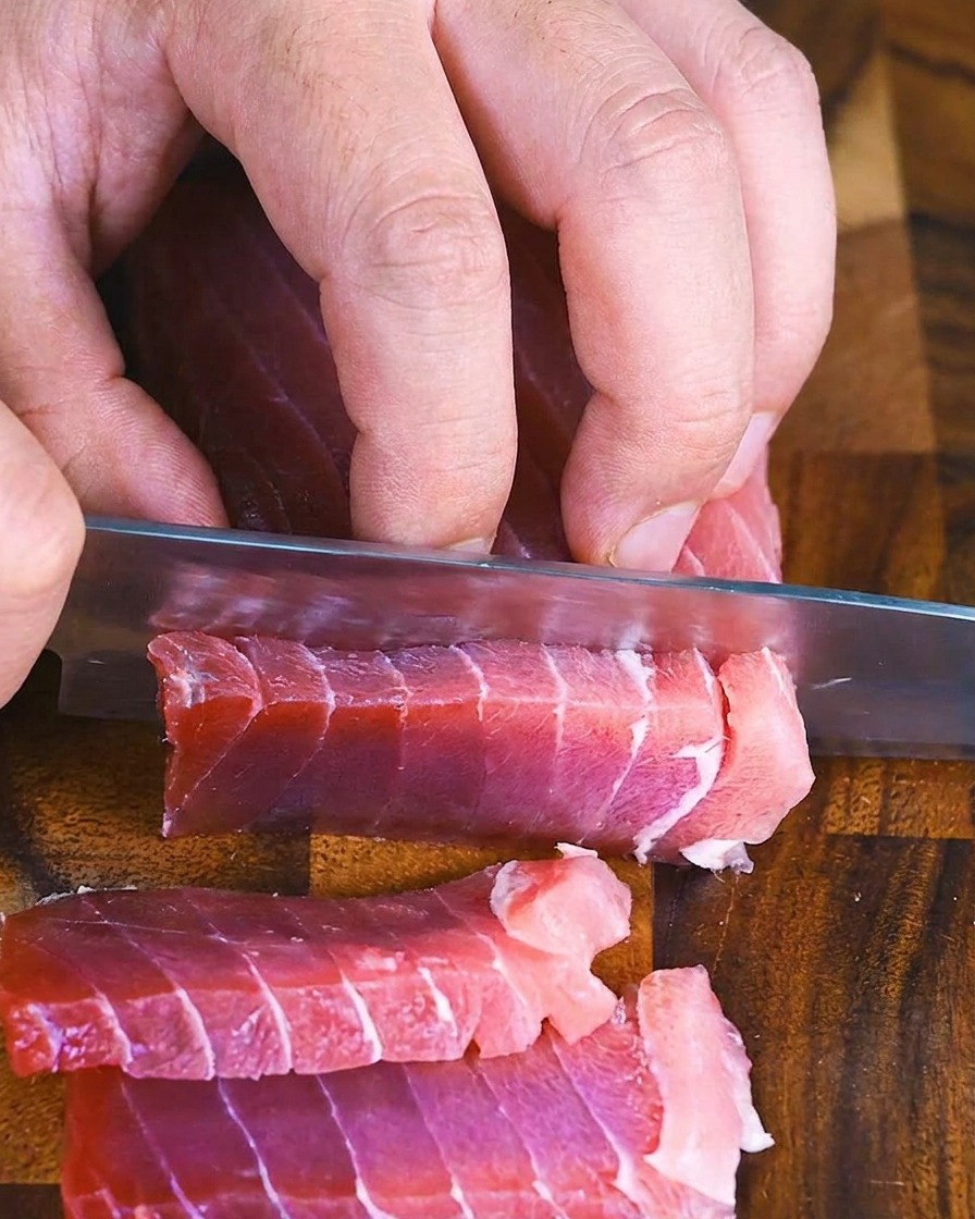Manos usando un cuchillo de chef para rebanar atún en rectángulos gruesos.