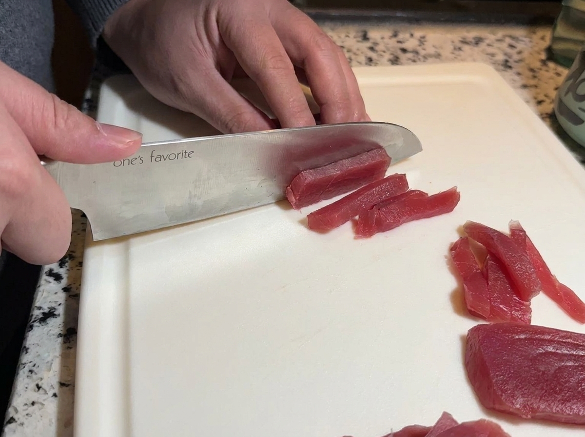Un cuchillo de chef cortando atún en tiras largas sobre una tabla.
