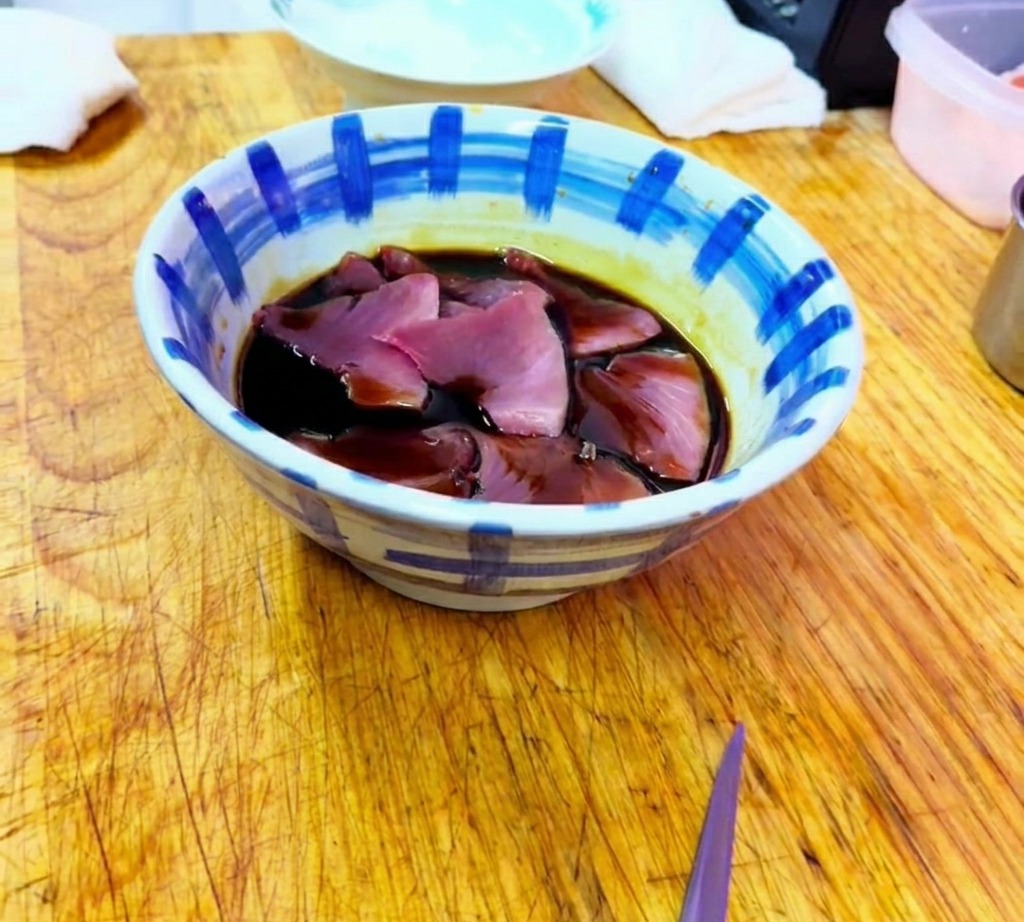 Láminas de atún crudo siendo sumergidas en un oscuro y sabroso adobo a base de soja dentro de un bol de cerámica estampada.