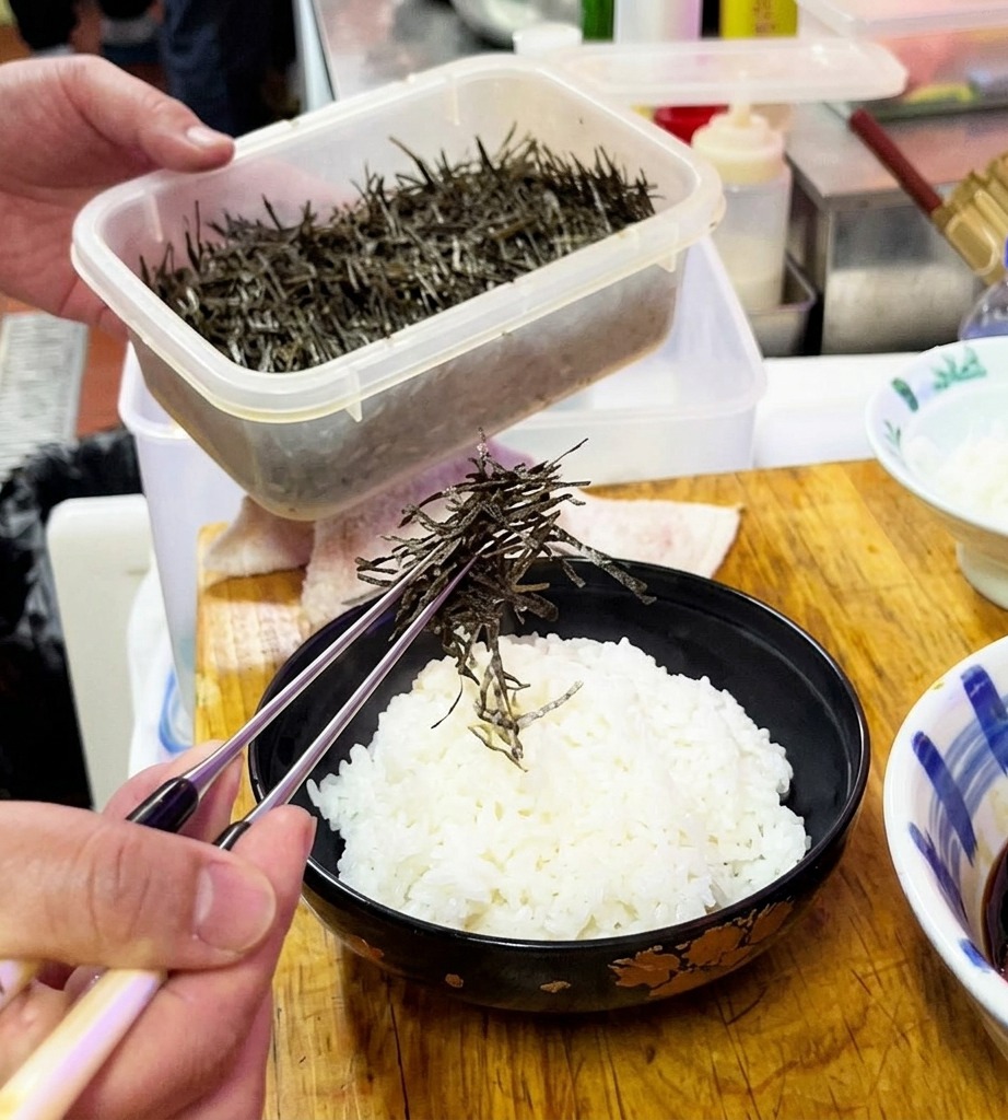 Algas secas en tiras siendo esparcidas sobre un bol de arroz blanco al vapor.