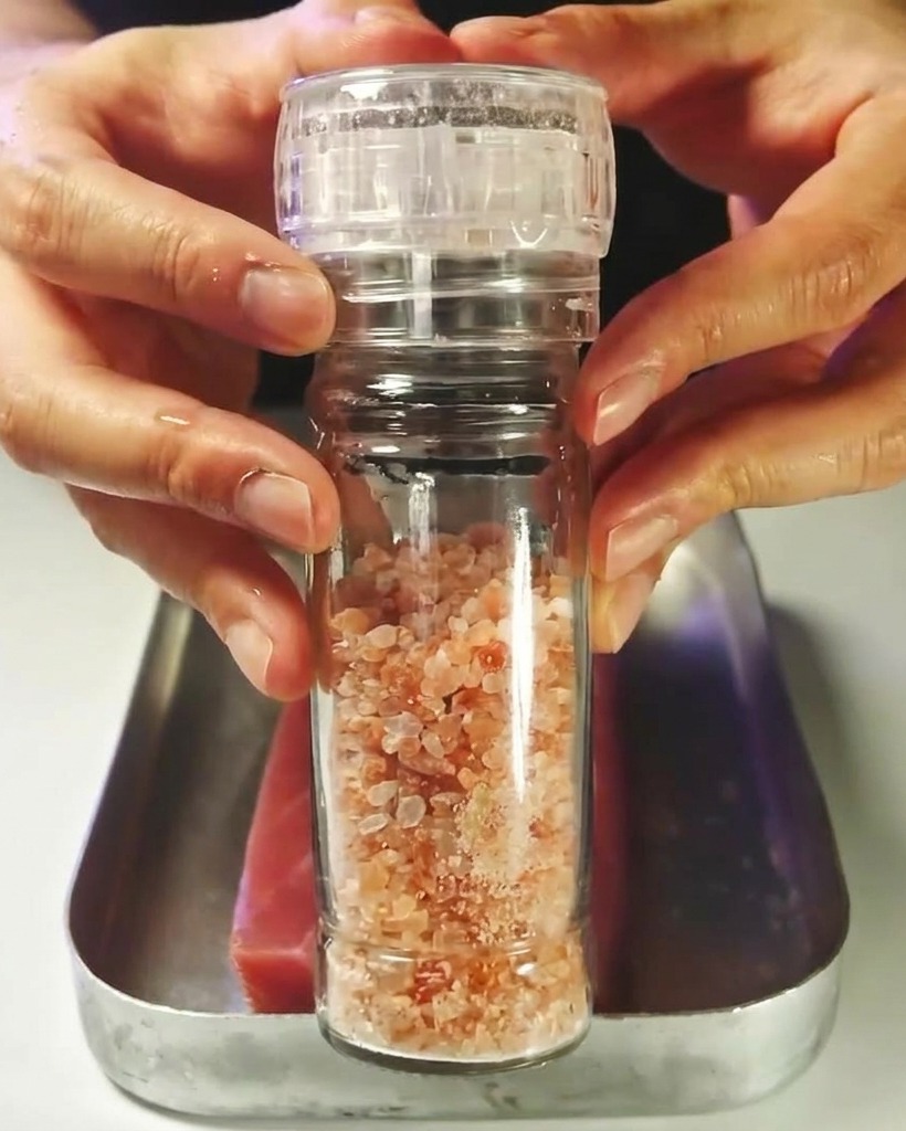 Un bloque de atún rojo crudo siendo sazonado con sal de un molinillo.