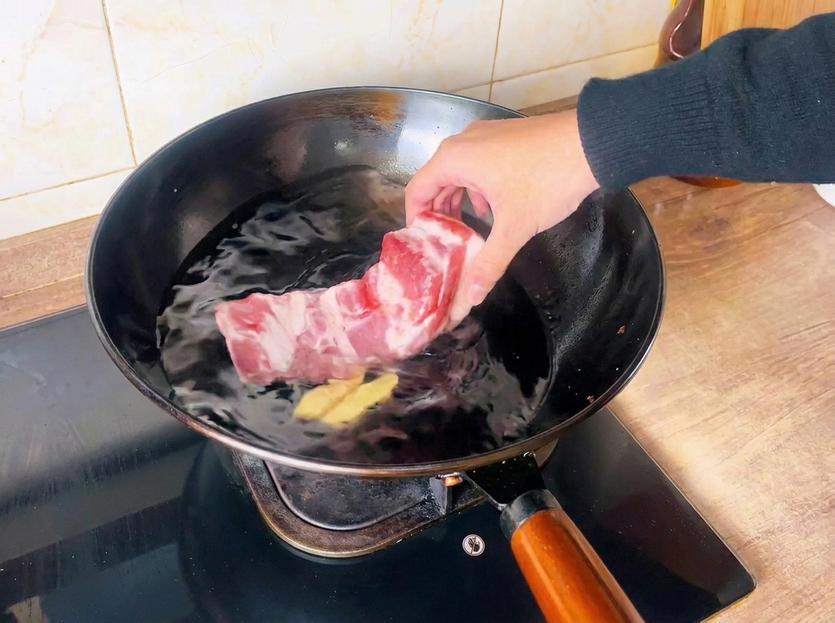 Una mano colocando un bloque de panceta de cerdo cruda en un wok con agua fría y rodajas de jengibre.