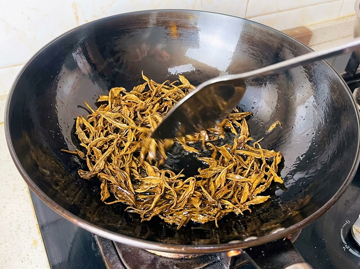 Hojas de té verde húmedas friéndose en aceite caliente dentro de un wok negro con una espátula de metal.
