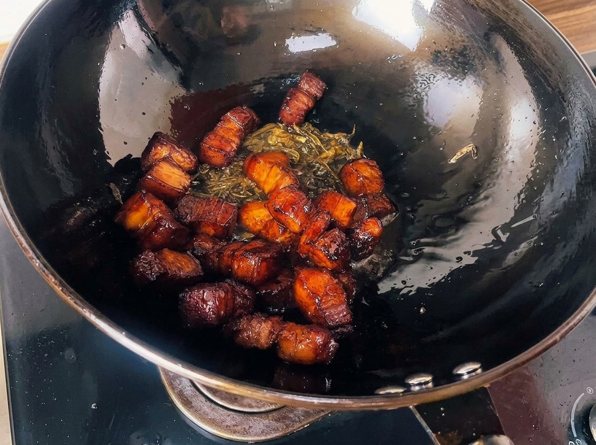 Cubos de panceta glaseada y asada mezclados con hojas de té fritas y crujientes, reposando dentro de un gran wok negro.