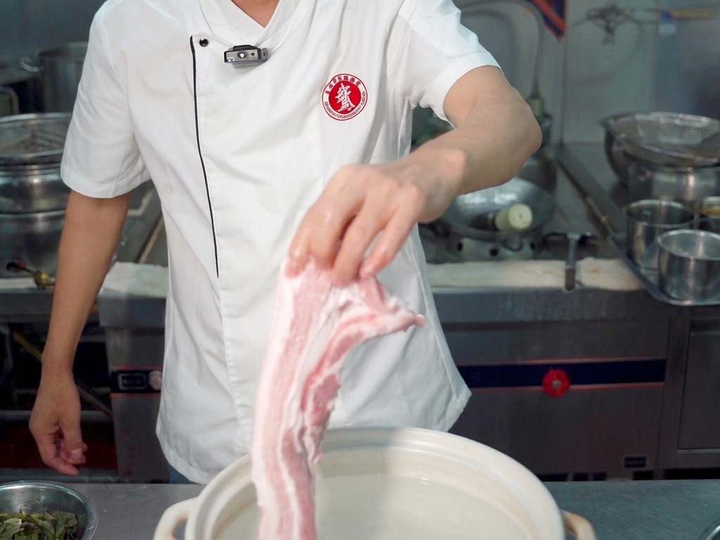 Un chef sosteniendo un trozo grande de panceta cruda sobre una olla con agua.