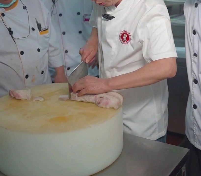 Un chef usando un cuchillo de carnicero para cortar un bloque de panceta hervida.