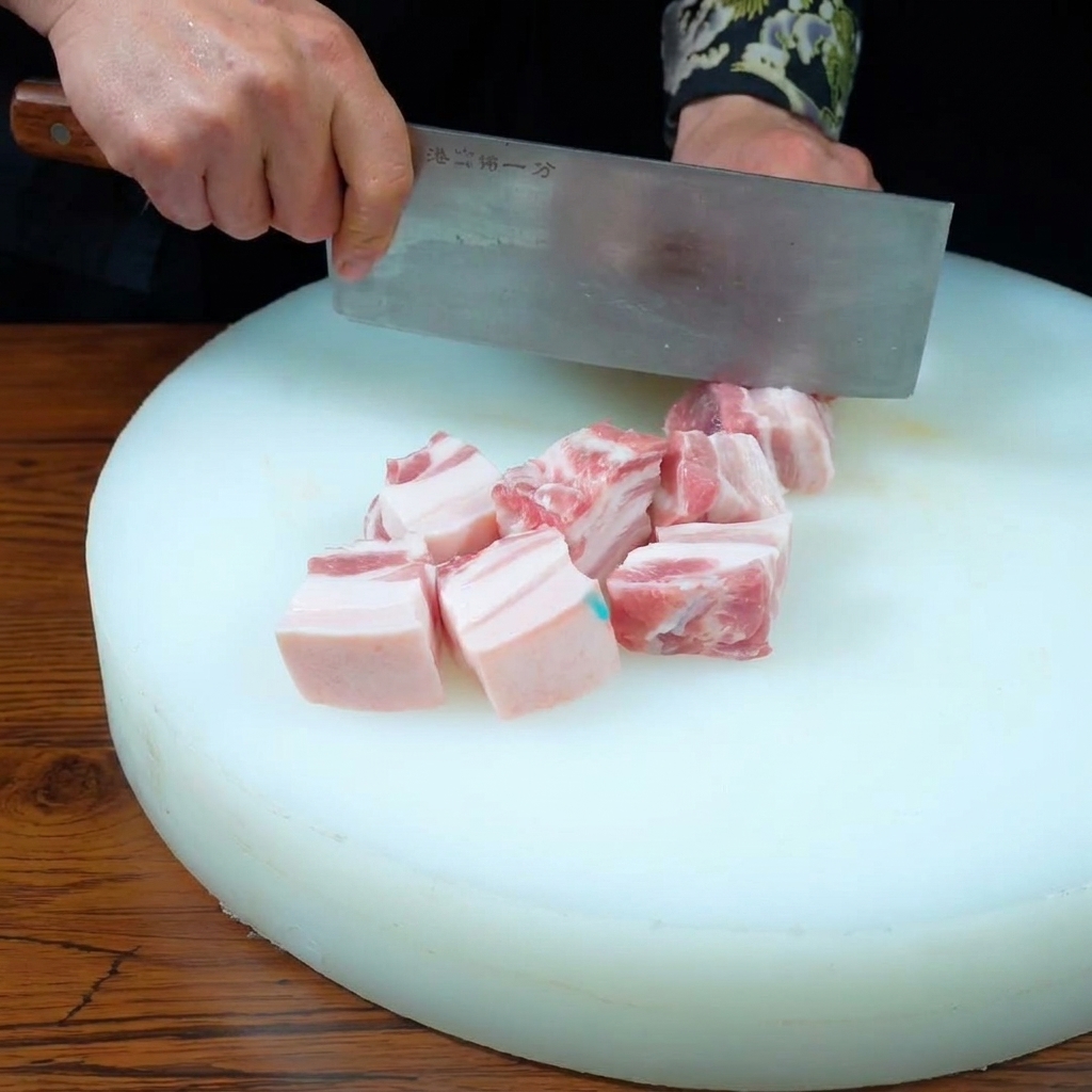 Manos usando un cuchillo de carnicero para cortar una pieza de panceta de tres capas en cubos uniformes sobre una tabla de cortar blanca.