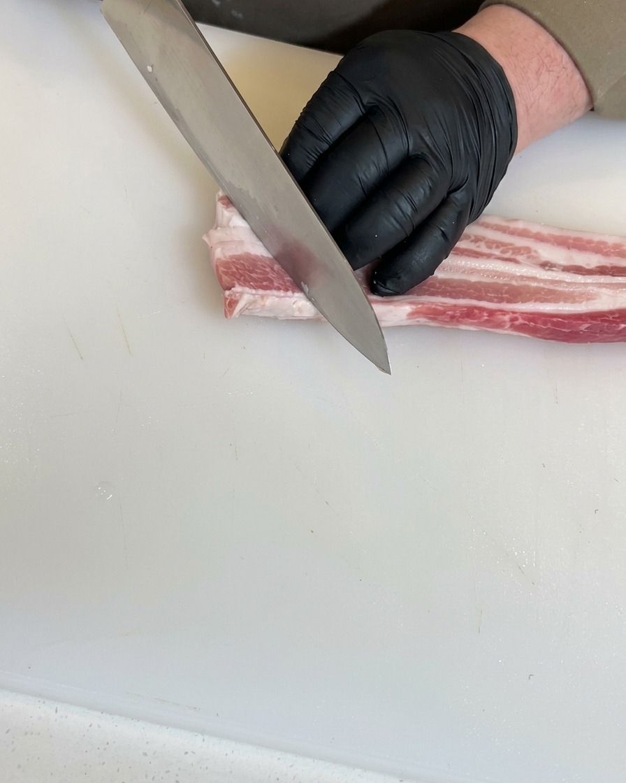 Manos con guantes negros usando un cuchillo de chef para hacer cortes diagonales profundos en una pieza de panceta cruda sobre una tabla de cortar blanca.