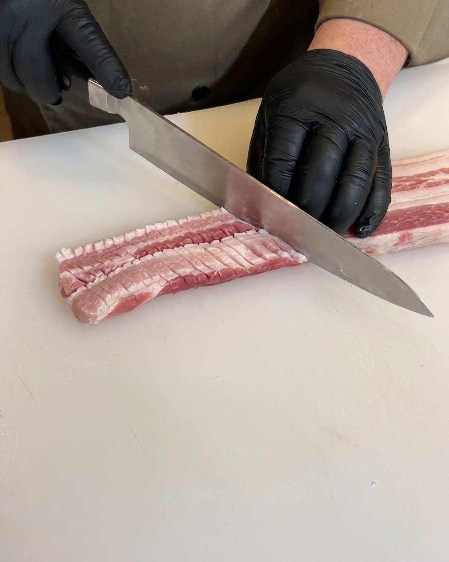 Manos con guantes negros marcando el lado inverso de una panceta de cerdo cruda con un cuchillo de chef, mostrando cortes profundos en cuadrícula.