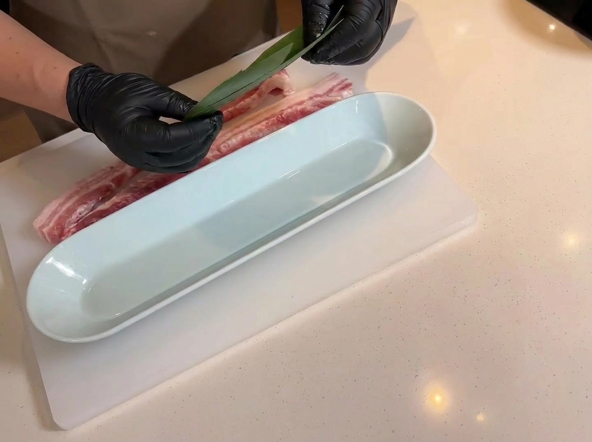 Manos sosteniendo una hoja de bambú verde sobre una bandeja blanca alargada, con la panceta cruda marcada cerca en la tabla de cortar.