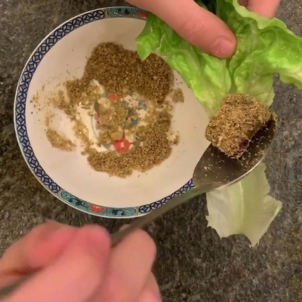 Primer plano de un trozo de panceta cocinada cubierta de cacahuetes triturados, a punto de colocarse sobre una hoja de lechuga.