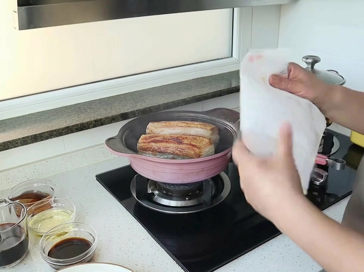 Manos sosteniendo una toalla de papel de cocina blanca sobre una sartén con dos trozos de panceta sellándose.