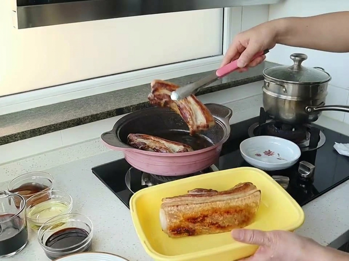 Pinzas levantando un trozo de panceta de cerdo bien dorado fuera de una sartén rosa.
