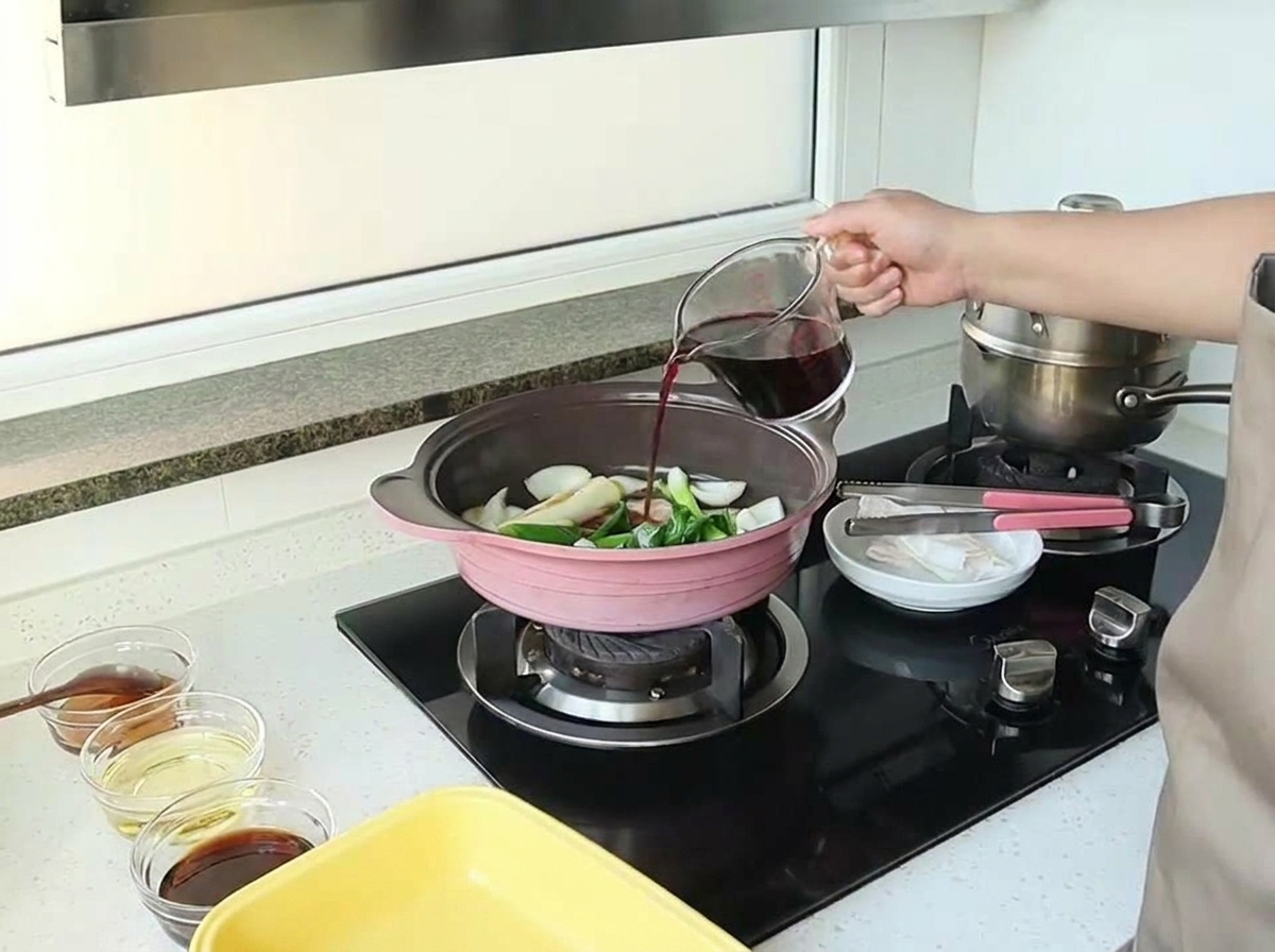 Una mano vertiendo vino tinto de una taza medidora de vidrio en una sartén con cebollas y aromáticos.