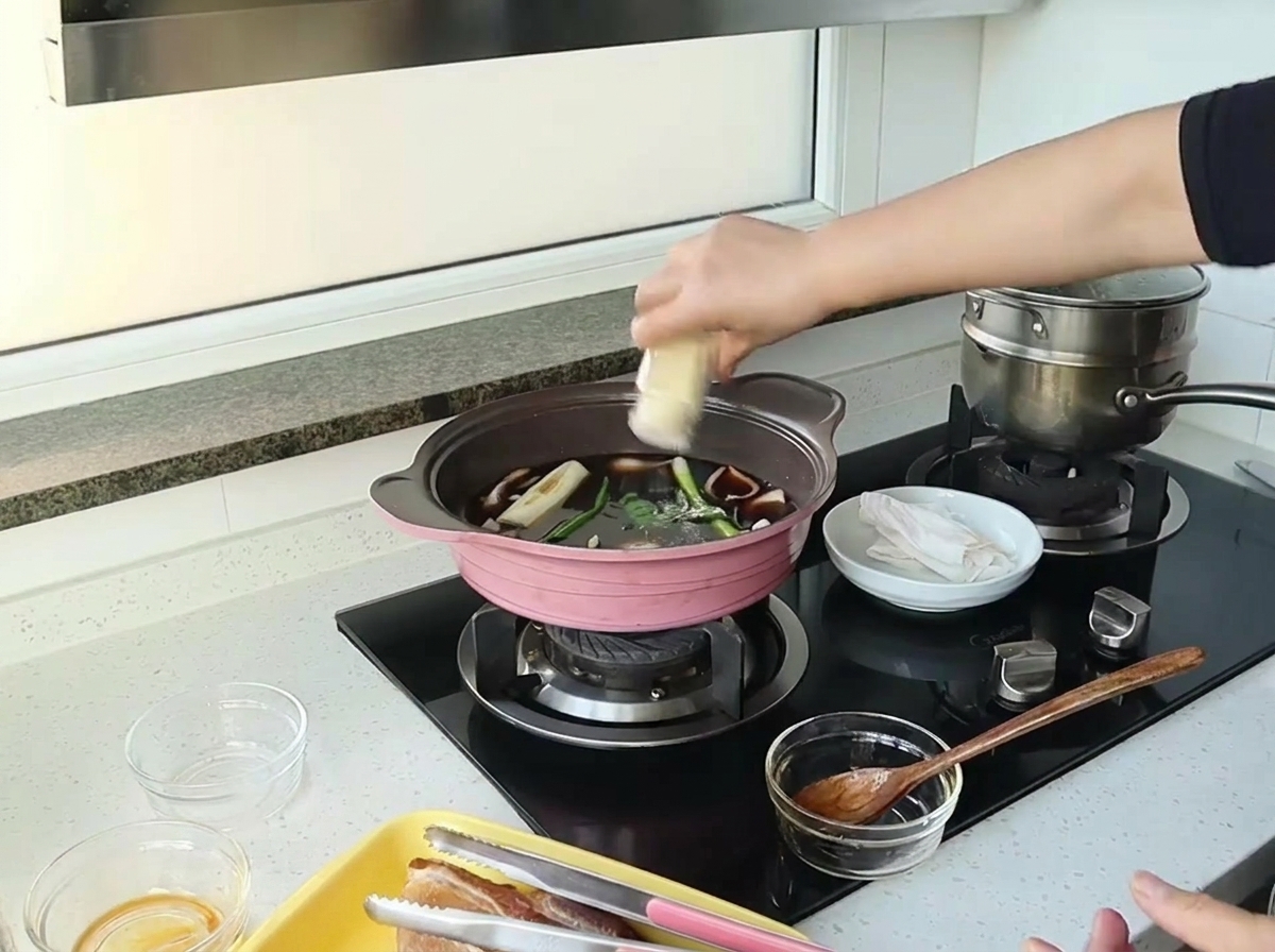 Una mano espolvoreando jengibre en polvo desde un frasco pequeño en una olla con salsa oscura, cebollas y cebolletas.