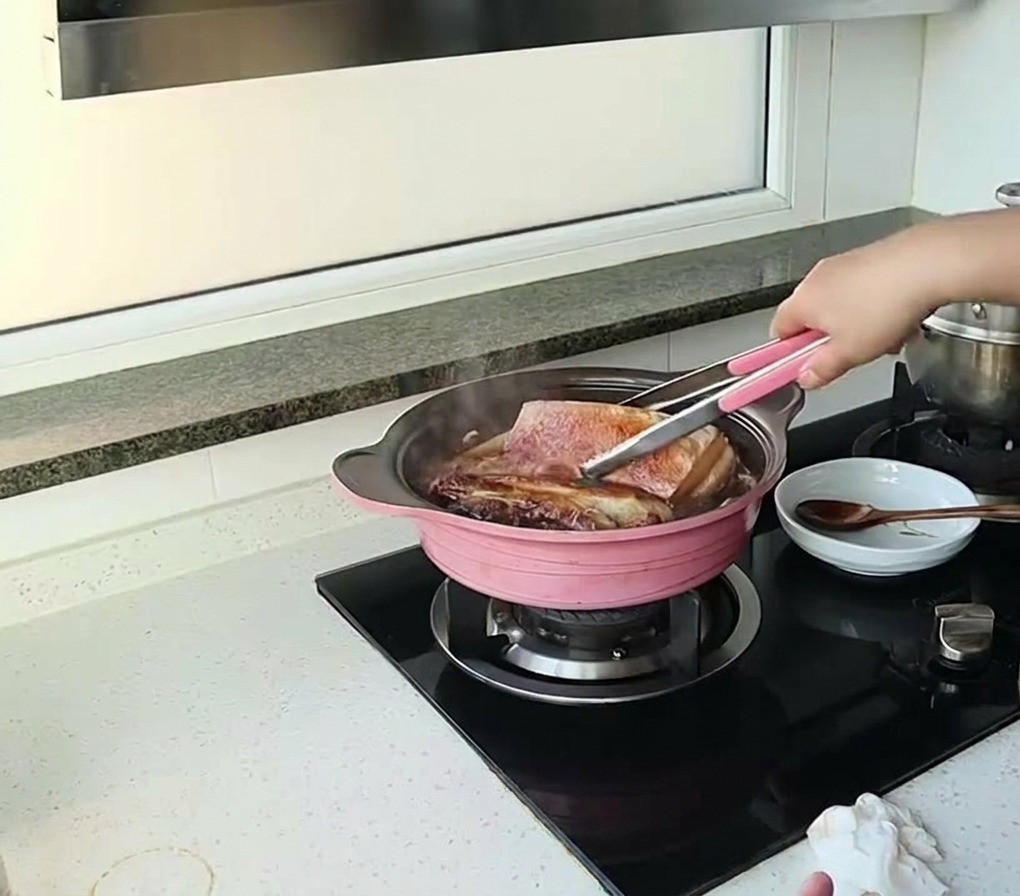 Una persona usando pinzas para voltear trozos grandes de panceta de cerdo cocinándose a fuego lento en una salsa oscura en una olla rosa.