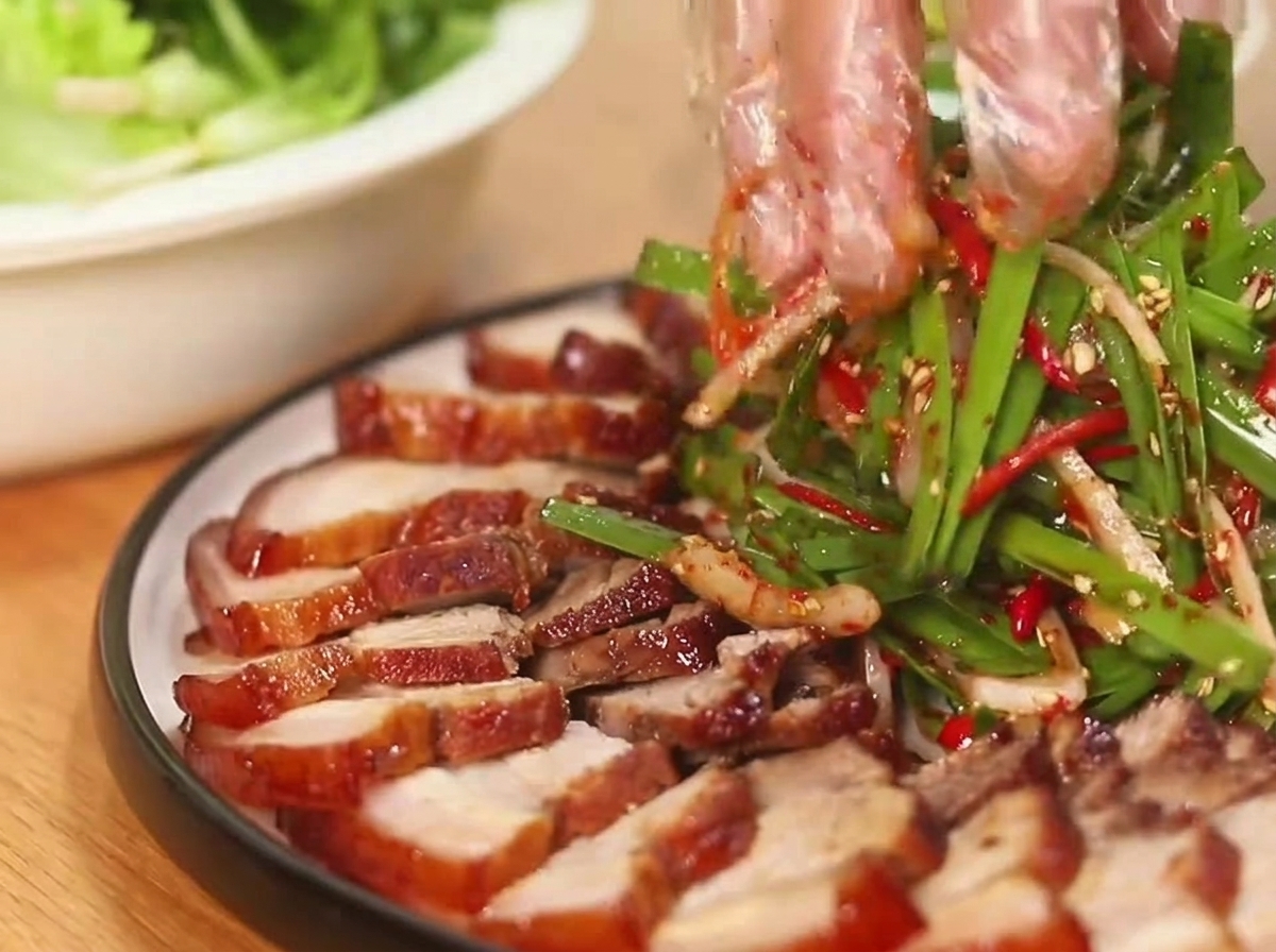 Manos enguantadas mezclando una ensalada de cebolleta verde vibrante y chile rojo junto a panceta de cerdo glaseada cortada dispuesta en un plato redondo.