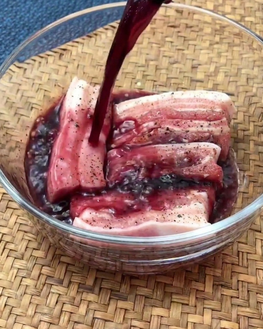 Trozos crudos de panceta marinando en un bol de cristal con vino tinto.