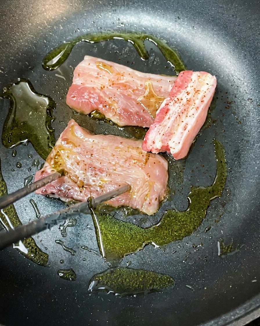 Trozos de panceta marinada dorándose en una sartén caliente con aceite de oliva.