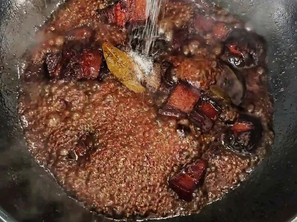 Sal espolvoreándose sobre los dados de panceta a fuego lento en una salsa rica y oscura.