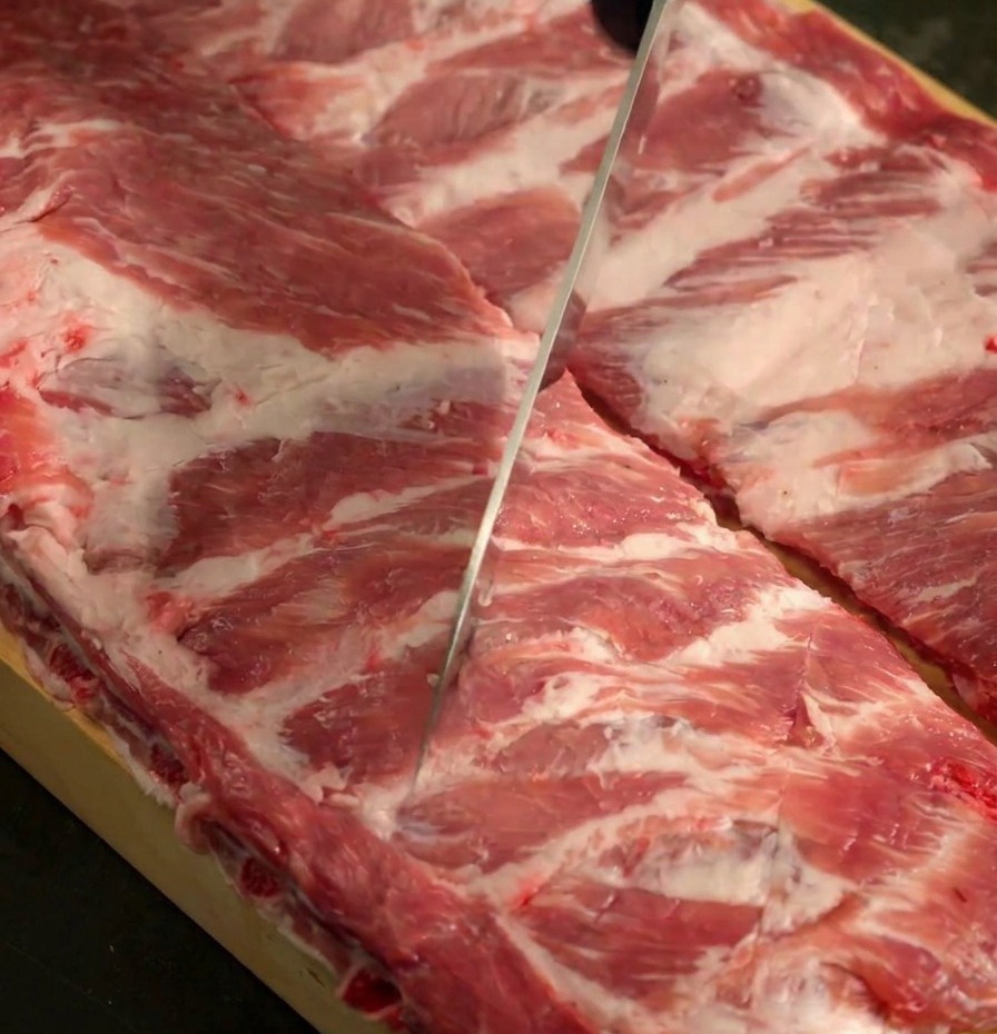 Un cuchillo afilado marcando la superficie de dos costillares de cerdo crudos sobre una tabla de madera.
