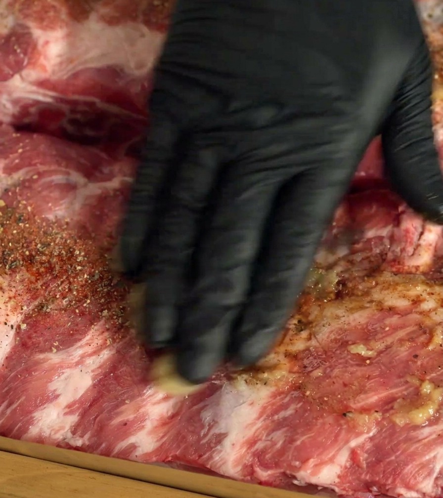 Una mano con guante frotando especias y ajo en las costillas de cerdo crudas.