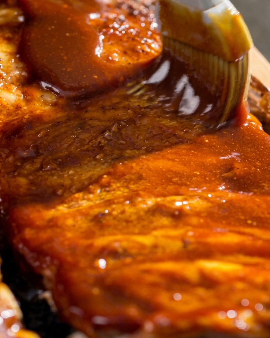 Primer plano de un pincel de cocina extendiendo un glaseado de barbacoa espeso y brillante sobre las costillas.