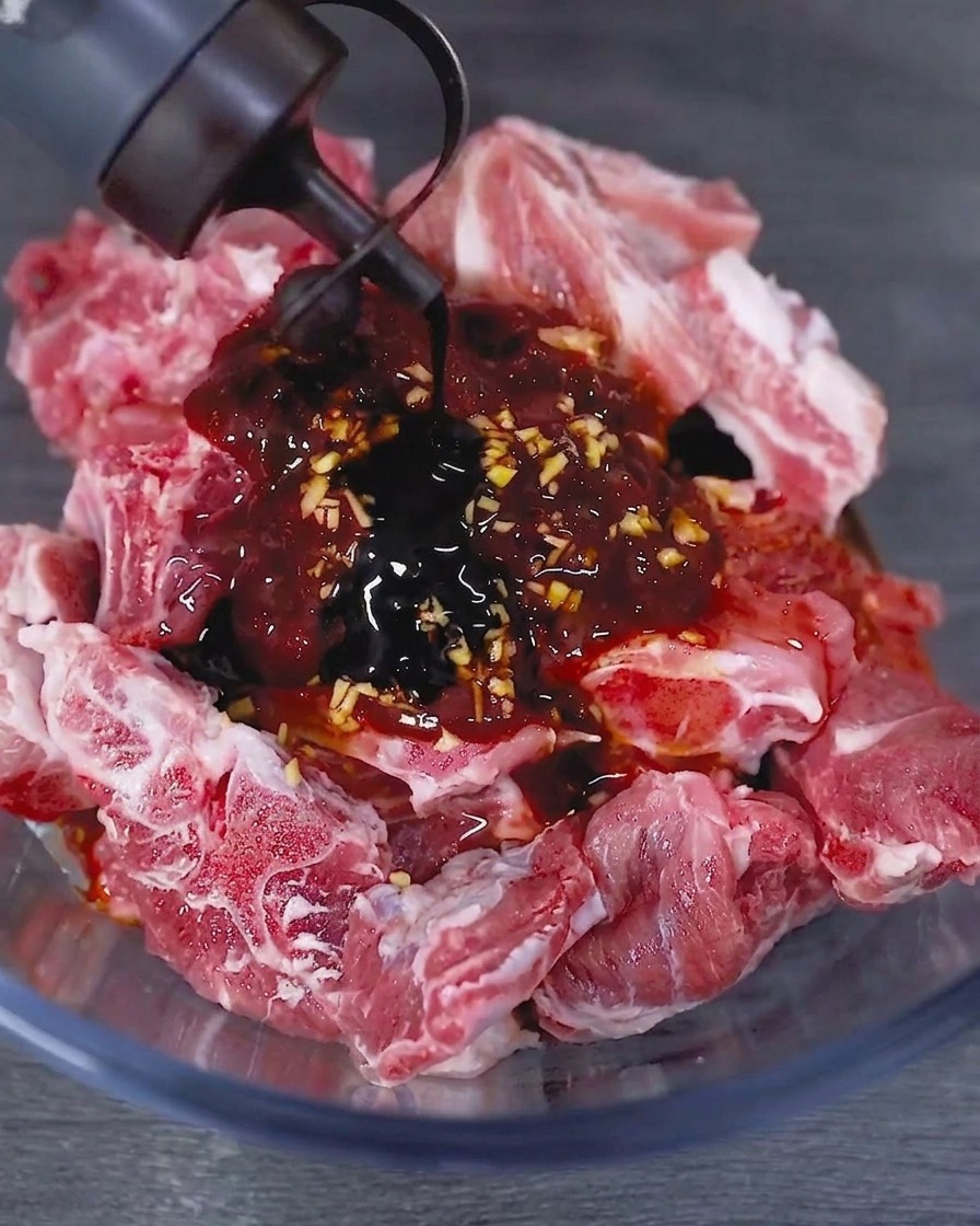 Salsa de soja oscura vertiéndose desde una botella a un tazón de vidrio que contiene costillas de cerdo crudas marinadas.