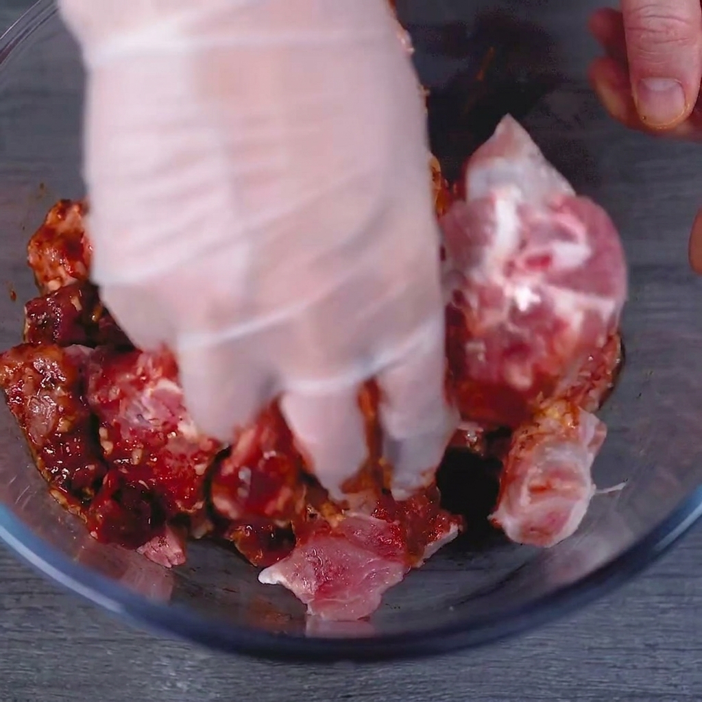 Una mano con guante masajeando a fondo la marinada roja en las costillas de cerdo crudas en un tazón de vidrio.