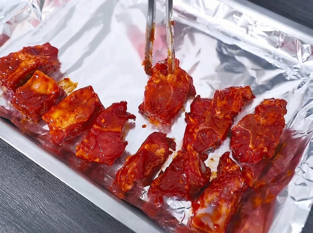 Costillas de cerdo marinadas crudas siendo dispuestas cuidadosamente en una bandeja para hornear forrada con papel aluminio usando pinzas de metal.