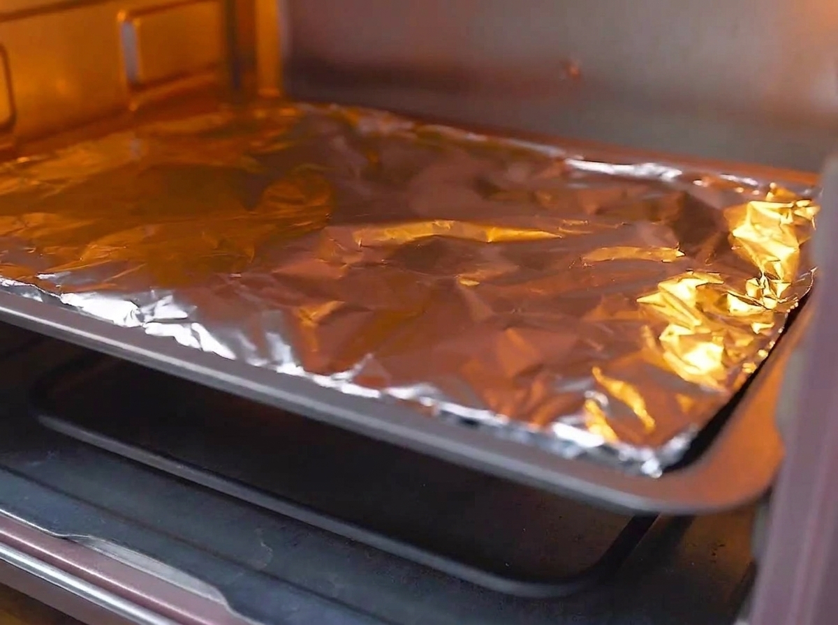 Una bandeja para hornear cubierta con papel aluminio siendo colocada en la rejilla central de un horno precalentado y brillante.