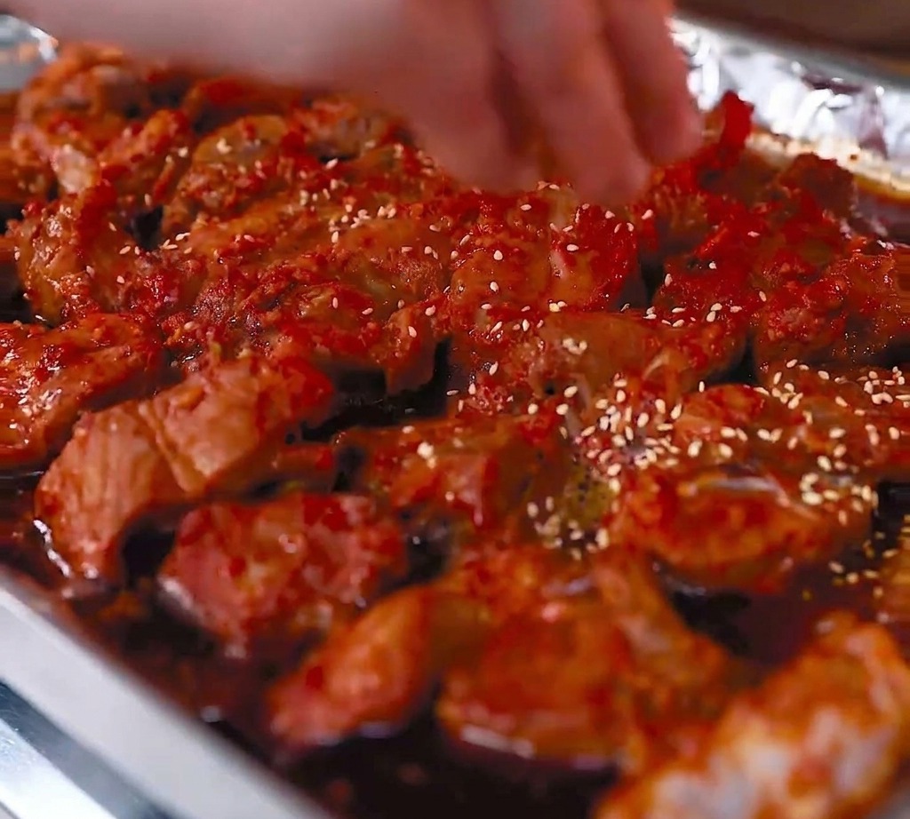 Una vista de primer plano de costillas de cerdo asadas en una bandeja forrada con papel aluminio, con semillas de sésamo blanco siendo espolvoreadas sobre la superficie brillante y glaseada de rojo.