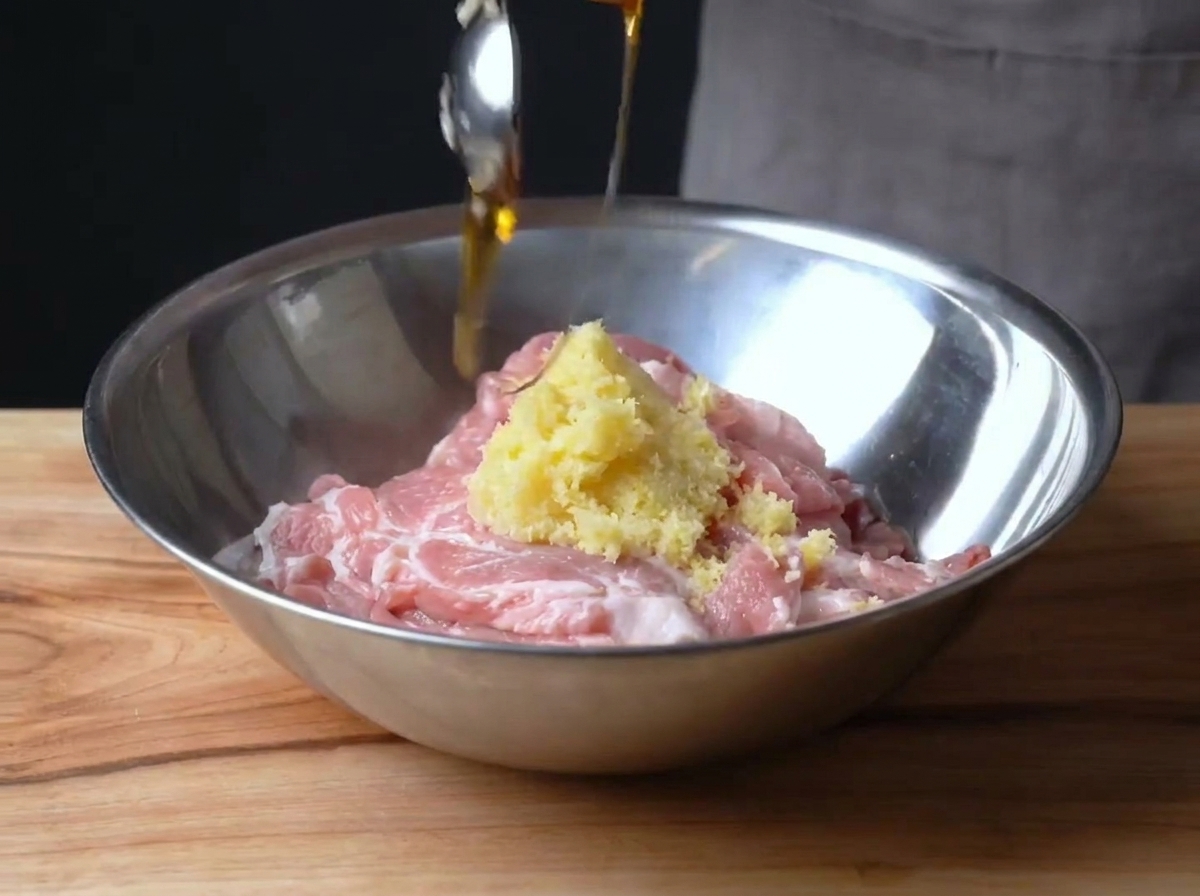 Cerdo crudo en láminas en un bol metálico siendo mezclado con ralladura de limón y jarabe de maíz.