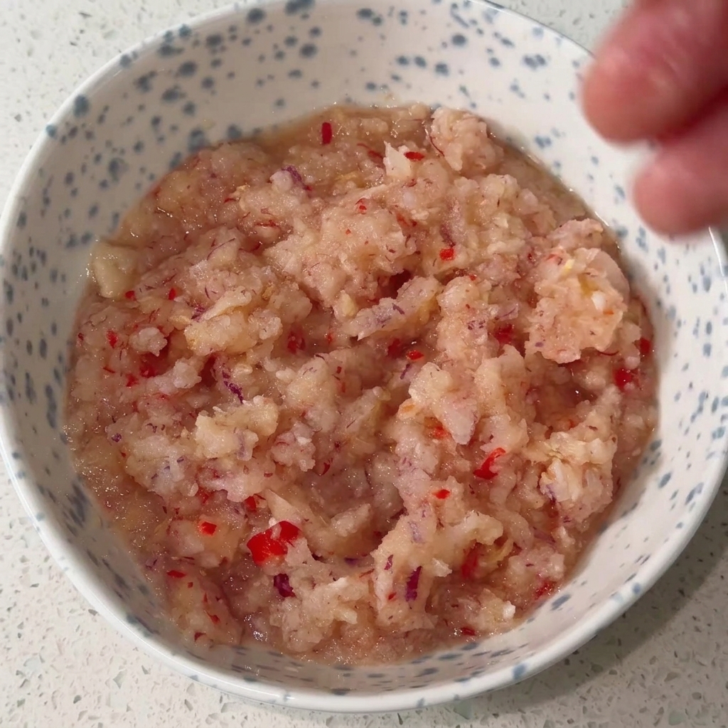 Un bol con la base de cebolla y chile siendo mezclada con condimentos líquidos y en polvo para crear una salsa roja rica.
