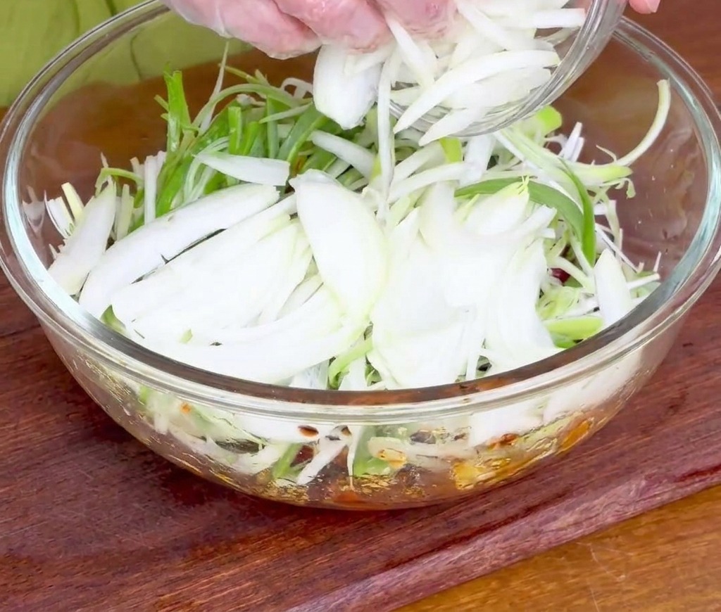 Manos añadiendo un puñado de cebolleta y cebolla blanca rebanada a la carne cruda marinada.