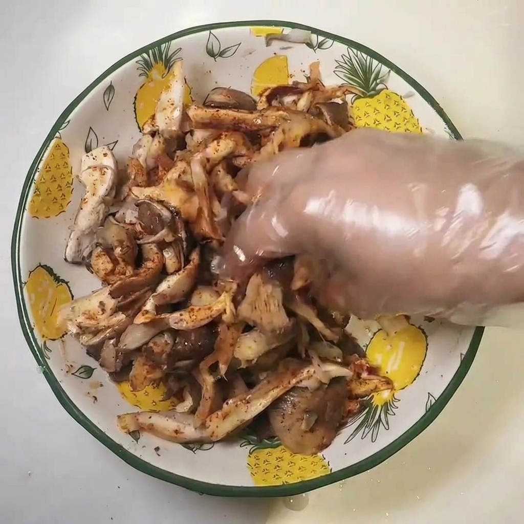 Una mano con guante de plástico mezclando setas sazonadas en un bol.