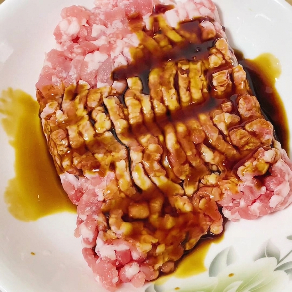 Salsa de soja siendo vertida sobre el cerdo picado crudo en un bol blanco.