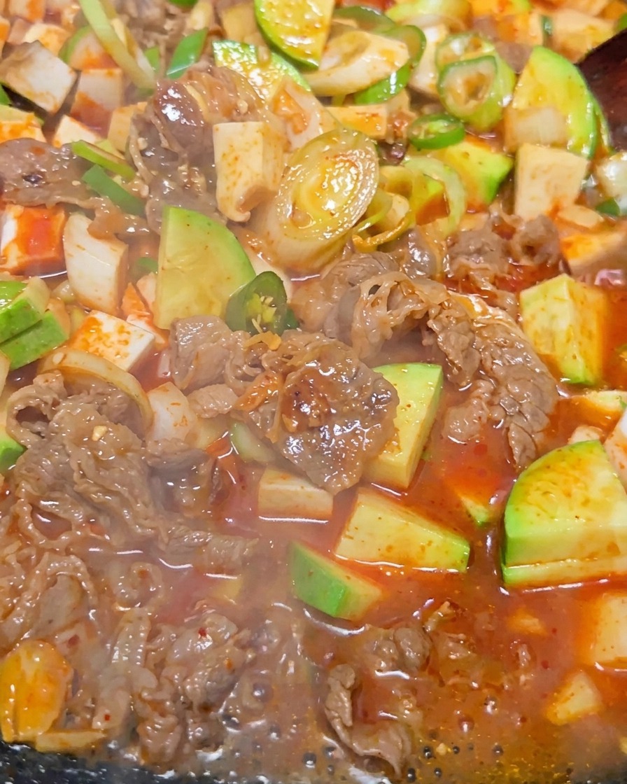 Primer plano del estofado coreano de pasta de soja burbujeando, mostrando trozos de tofu, ternera y calabacín verde en un caldo marrón rojizo.